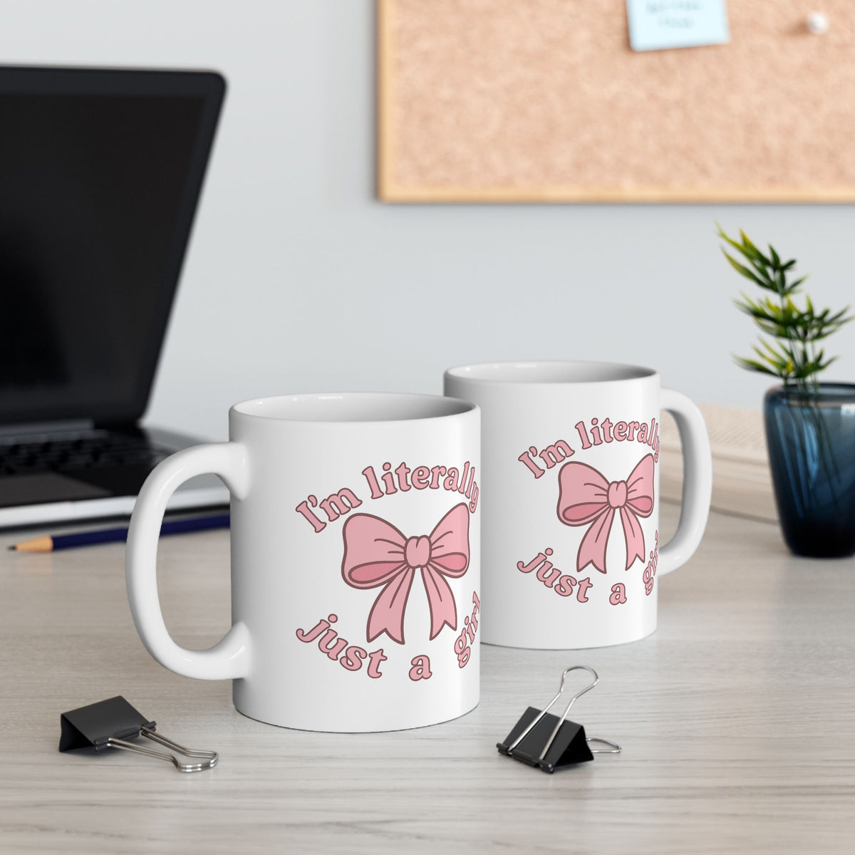 Two white mugs with pink bows and text on a desk with office supplies.