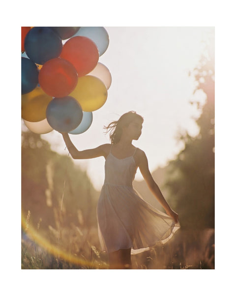 whimsical photography poster of a young woman holding colorful balloons in a field with a warm glow