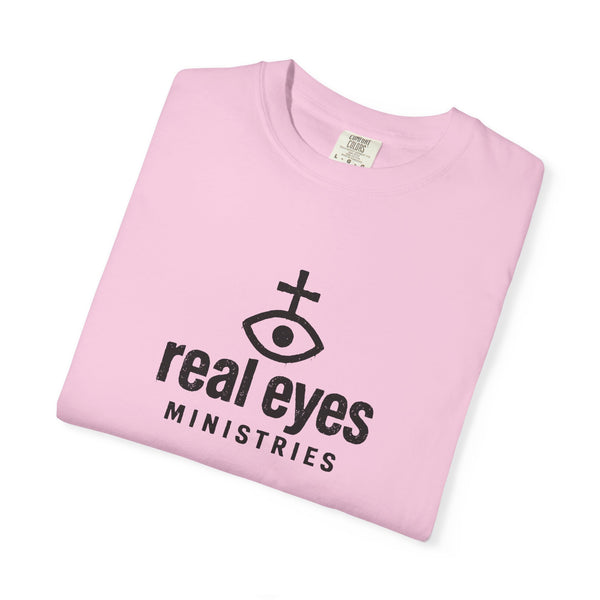 Folded pink t-shirt with 'real eyes MINISTRIES' logo on a white background