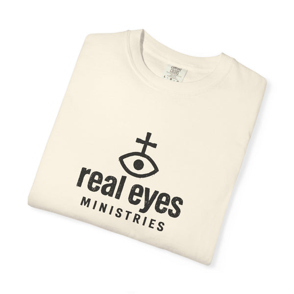 Folded cream-colored t-shirt with 'real eyes MINISTRIES' text and logo on a white background