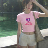 Person wearing a pink t-shirt with 'LOVE SPACES' text and a heart graphic, standing outdoors by a body of water.