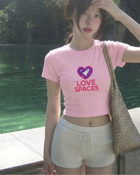 Person wearing a pink t-shirt with 'LOVE SPACES' text and a heart graphic, standing outdoors by a body of water.