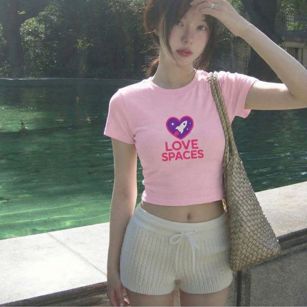 Person wearing a pink t-shirt with 'LOVE SPACES' text and a heart graphic, standing outdoors by a body of water.