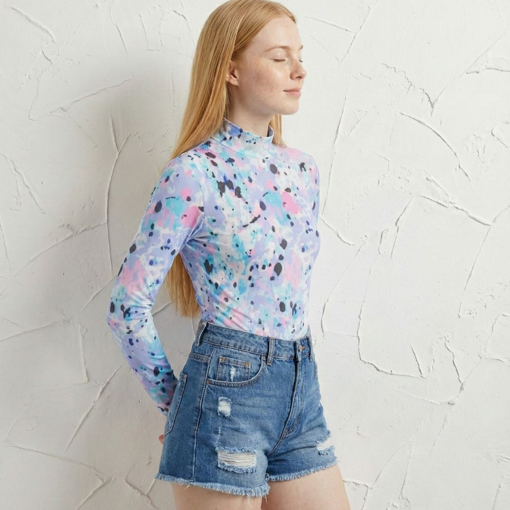 Woman wearing a colorful long-sleeve top and denim shorts against a textured white wall.