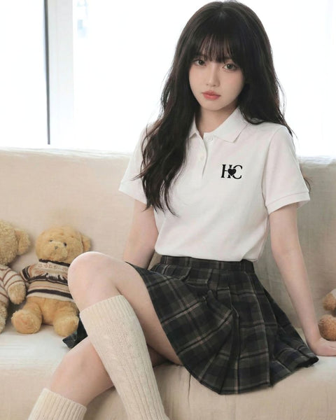 Woman in a white polo shirt with a Heart Circle logo and plaid skirt sitting on a couch with teddy bears.