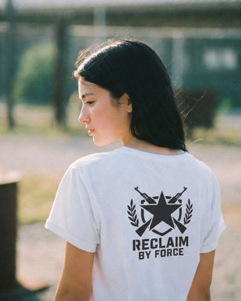 Person wearing a white t-shirt with a military design and "reclaim by force" text on the back, standing outdoors.
