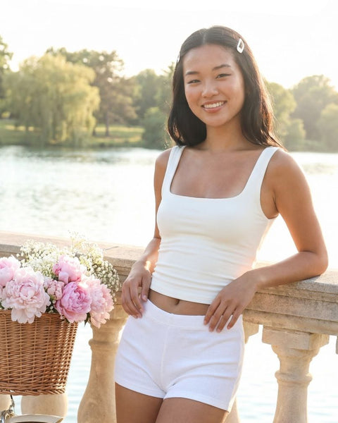 Woman in white tank top and shorts standing by a lake with a bicycle and flowers in the background