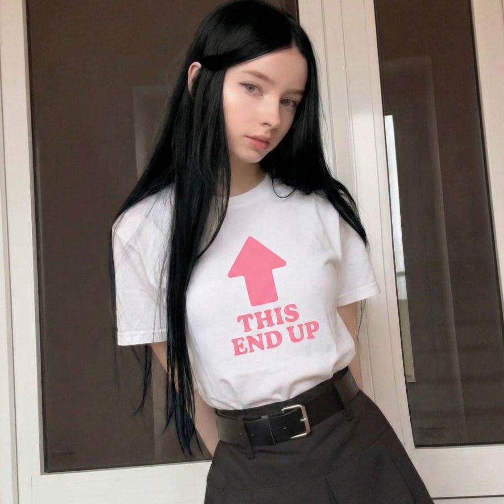 Woman wearing a white t-shirt with a pink graphic and text that reads "this end up," standing indoors.