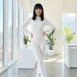 Asian woman wearing a white long sleeve jumpsuit in a bright, modern room with large windows and plants.