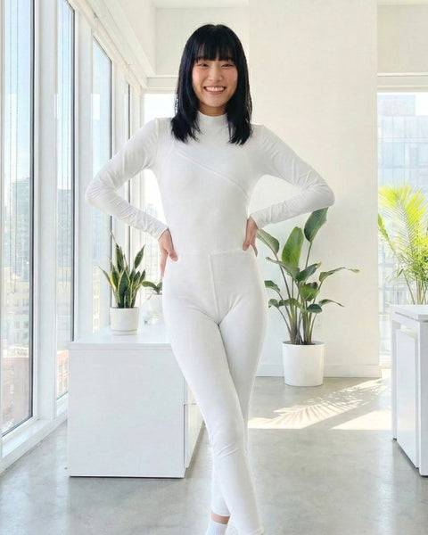 Asian woman wearing a white long sleeve jumpsuit in a bright, modern room with large windows and plants.