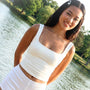 Woman in a white square neck tank top standing by a body of water with greenery in the background