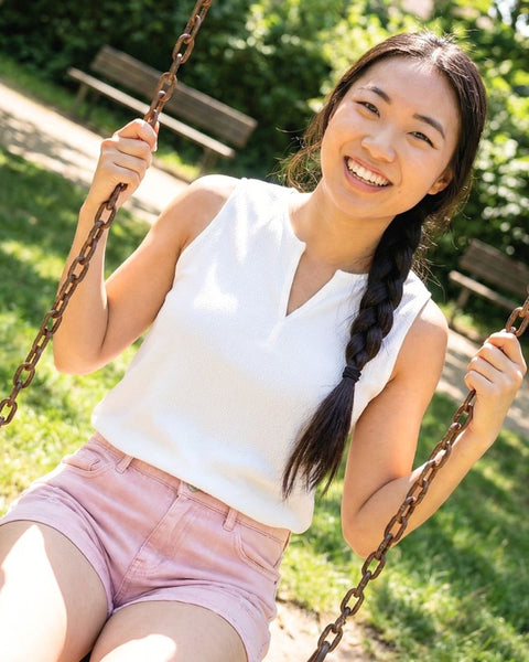 Woman wearing a white v-neck sleeveless top and pink shorts swinging on a swing set in a park with greenery